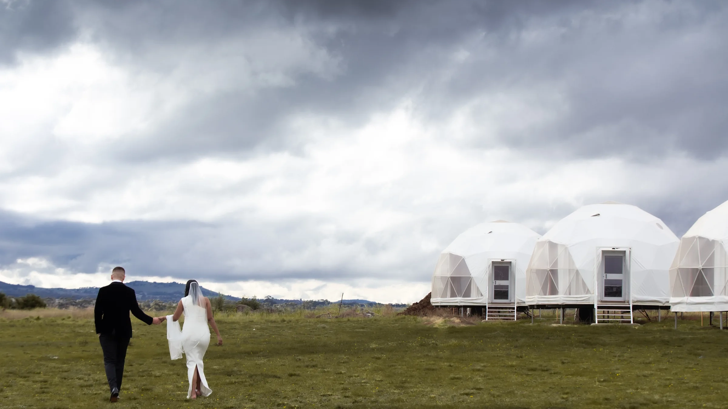 A bride and groom walk hand in hand across an open field toward geodesic dome tents beneath a dramatic storm sky, photographed by Royale Photography in regional Victoria.