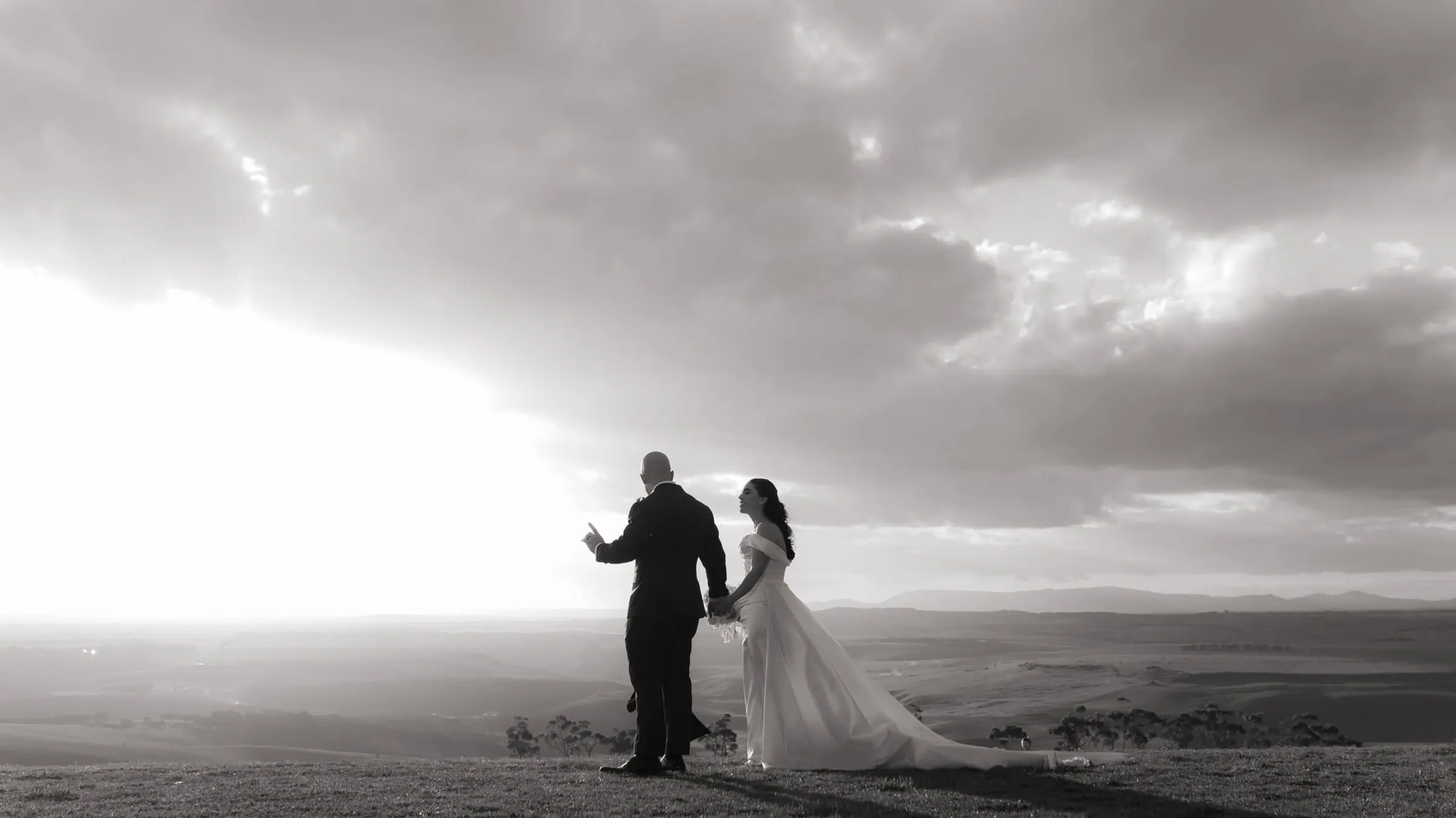 Black-and-white wedding portrait of a bride and groom holding hands beneath dramatic clouds, photographed by Royale Photography.