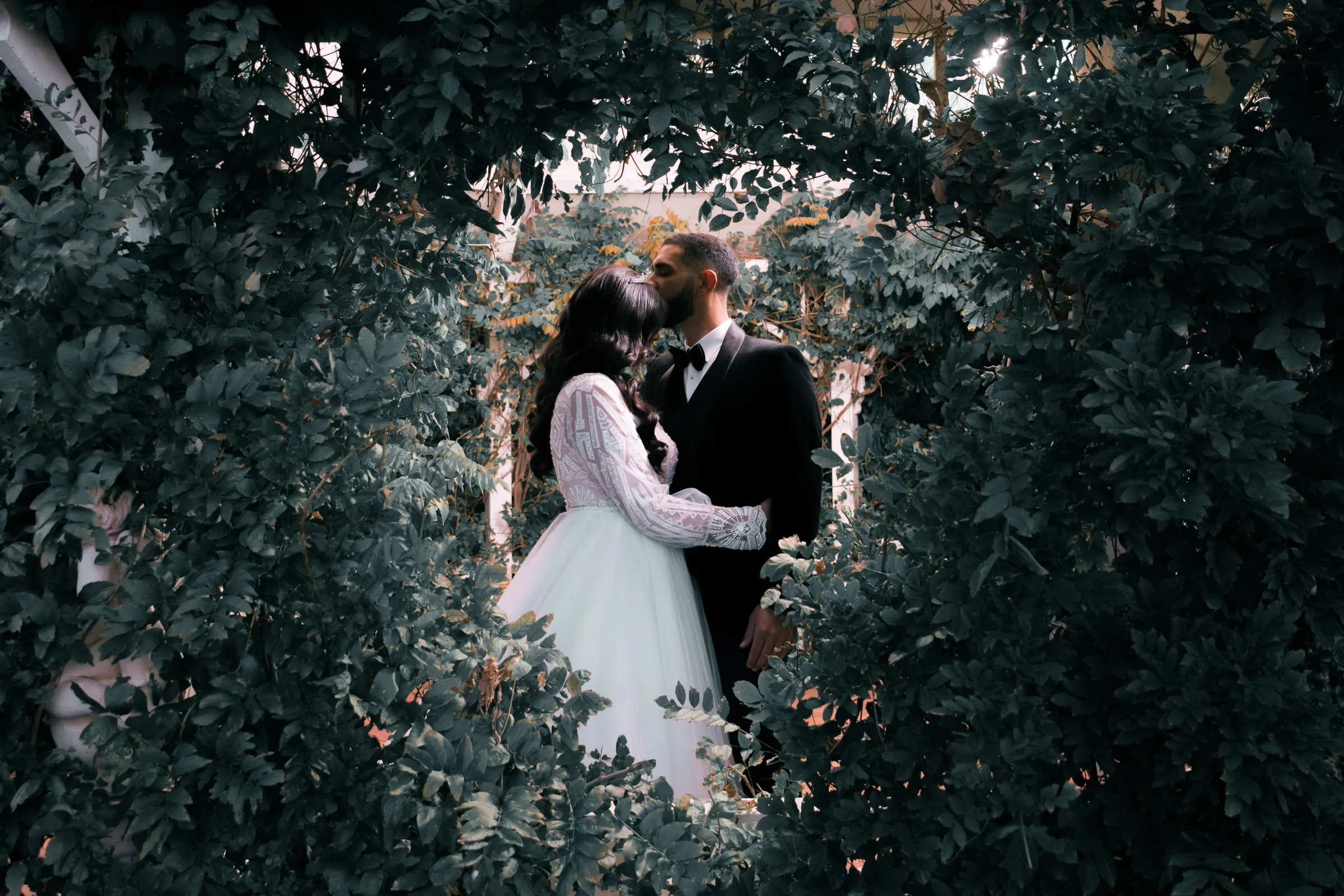 A couple embrace framed by lush green foliage on their wedding day, photographed by Royale Photography.